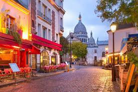A cobblestone street in Montmartre with a cafe as well as Sacre-Caeur basilica in the background.